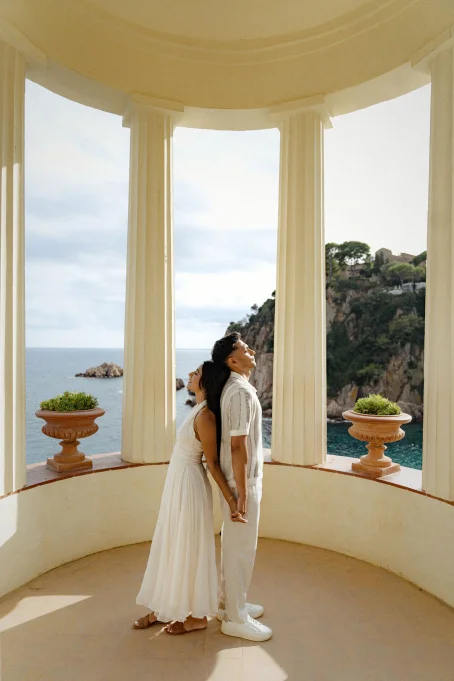 Romantic couple holding hands and leaning back to back, framed by Marimurtra garden’s seaside pavilion.