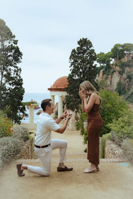 Romantic proposal at Marimurtra garden, man on one knee as woman reacts with joy, captured by a professional photographer.
