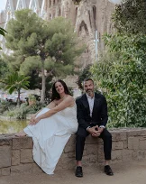 Smiling couple in wedding attire sits by lush greenery at La Sagrada Familia, captured by a professional photographer.