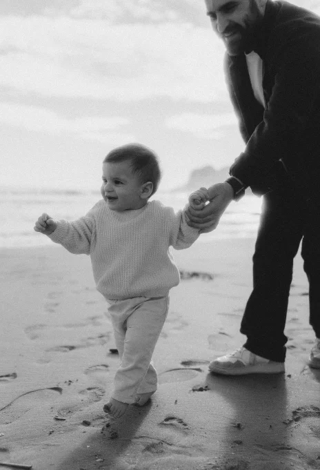 Smiling toddler takes first steps on Ginesta beach, guided by a parent during a family photography session.