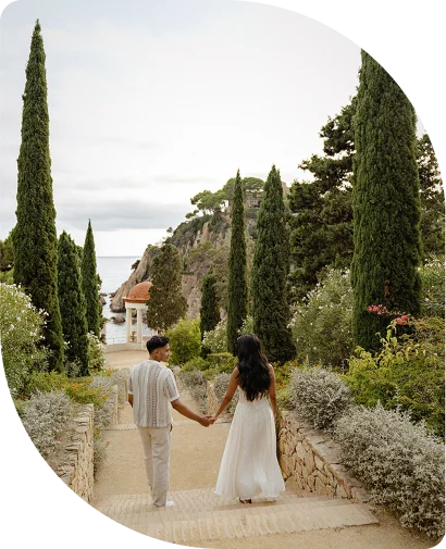 Couple holding hands during a surprise proposal photoshoot in a scenic Barcelona garden, walking toward the sea.
