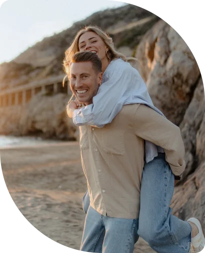Laughing couple enjoys a playful piggyback ride on a Barcelona beach during an engagement photoshoot.