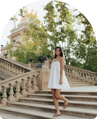 Smiling woman in a white dress poses on grand stone steps during a Vacation Portraits photoshoot in Barcelona.