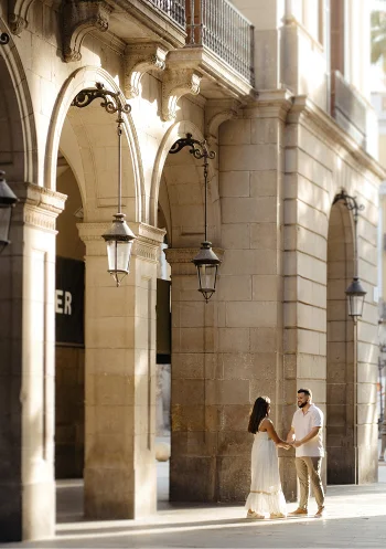 A couple holds hands under sunlit arches in a romantic engagement session in Barcelona’s historic city center.