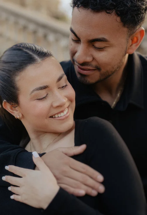 A couple shares a warm embrace outdoors, smiling and gazing at each other during an engagement session.