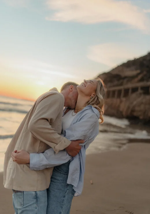 Couple laughing and embracing on a Barcelona beach at sunset, captured during a Vacation Portraits photoshoot.