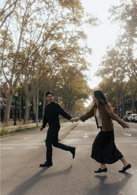 Couple laughing and holding hands while crossing a tree-lined street during a Vacation Portraits shoot in Barcelona.