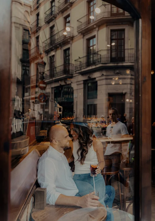 Couple sharing a joyful moment over drinks in a cozy Barcelona café, captured during an engagement photoshoot.