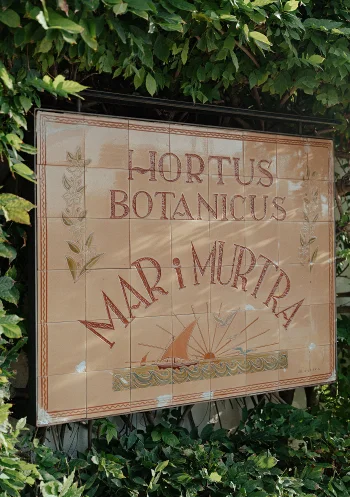 Decorative sign for Hortus Botanicus Mar i Murtra in Barcelona, framed by lush green leaves.