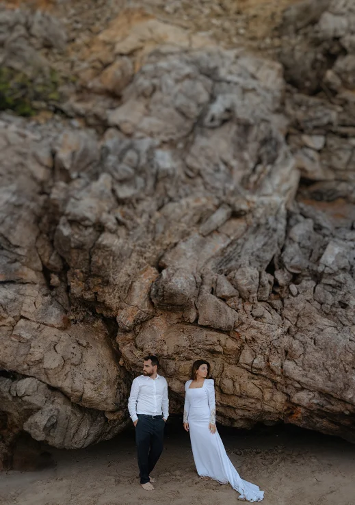 Engaged couple stands barefoot on a Barcelona beach, gazing apart against dramatic rocky cliffs.