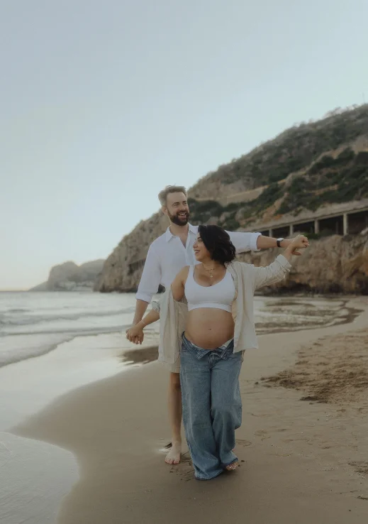 Joyful couple expecting a baby walk barefoot on a Barcelona beach, Vacation Portraits photoshoot capturing their happiness.