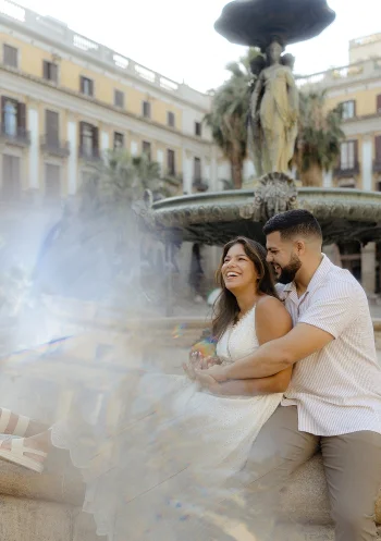 Joyful couple laughing together by a fountain in Barcelona, captured during an engagement photo session.