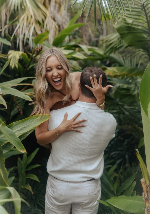 Joyful surprise proposal in lush Barcelona garden, woman laughing and showing engagement ring as partner lifts her.