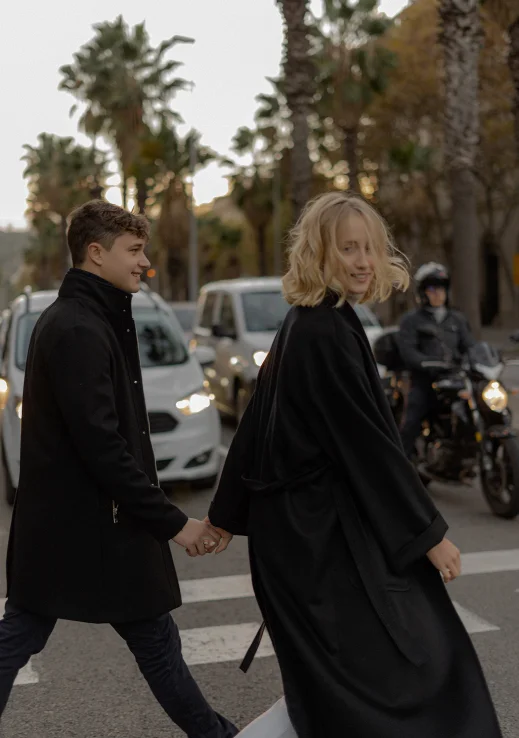 Smiling couple holding hands crossing a palm-lined street in Barcelona, captured during an engagement photoshoot.