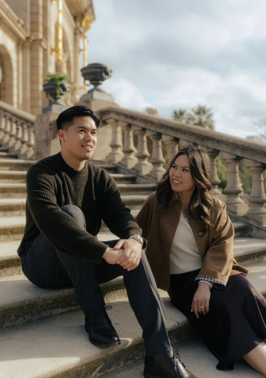 Smiling couple sitting on stone steps in Barcelona, sharing a candid moment during an engagement photoshoot.