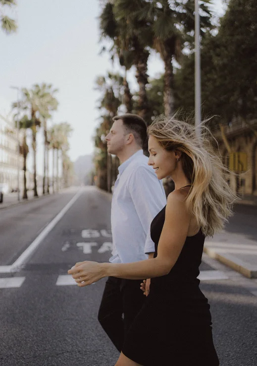 Smiling couple walking hand in hand down a palm-lined street in Barcelona during an engagement photoshoot.