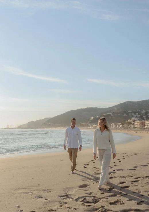 Smiling couple walks barefoot along a sunny Barcelona beach during an engagement photoshoot, mountains in the background.