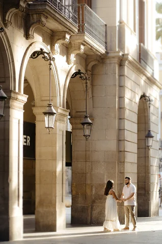 A couple holding hands and smiling under sunlit arches in Barcelona, captured during an engagement photoshoot.