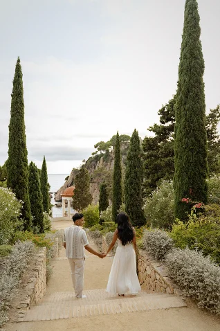 Couple holding hands during a surprise proposal photoshoot in a scenic Barcelona garden, walking toward the sea.