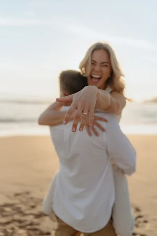 Joyful couple embraces on a Barcelona beach as she shows off her engagement ring after a surprise proposal photoshoot.