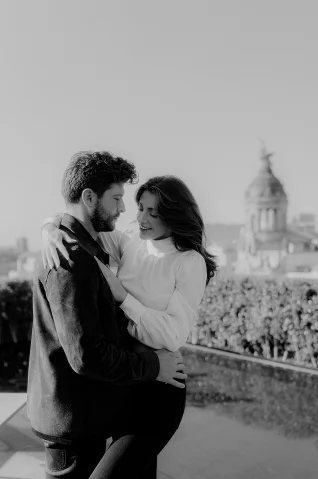 Smiling couple embraces during an engagement photoshoot in Barcelona, with city architecture in the background.