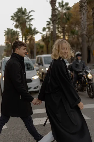 Smiling couple holding hands crossing a palm-lined street in Barcelona, captured during an engagement photoshoot.