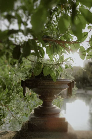 Soft morning light filters through lush leaves over a classic stone urn by a tranquil lake in Barcelona.