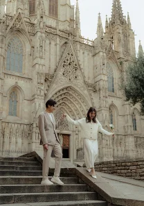 A couple holding hands and smiling as they walk down steps outside a cathedral, captured during an engagement photoshoot.
