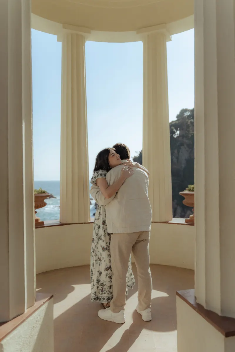 Couple embracing after a surprise proposal photoshoot by the sea, framed by elegant columns and natural light.