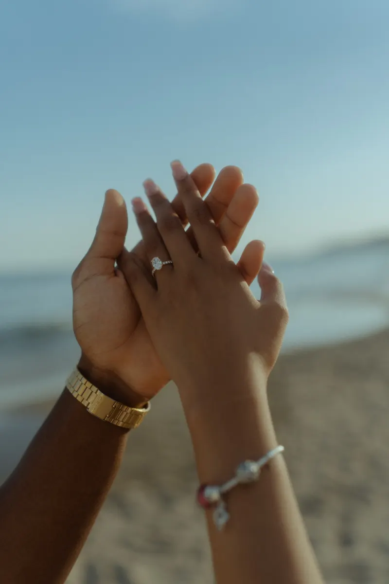 Couple holding hands on the beach, showing off an engagement ring during a surprise proposal photoshoot.