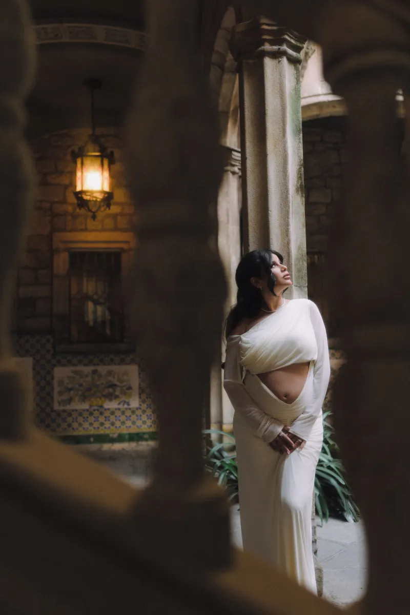 Expectant mother in elegant white dress poses serenely during a vacation portraits photoshoot in a historic courtyard.