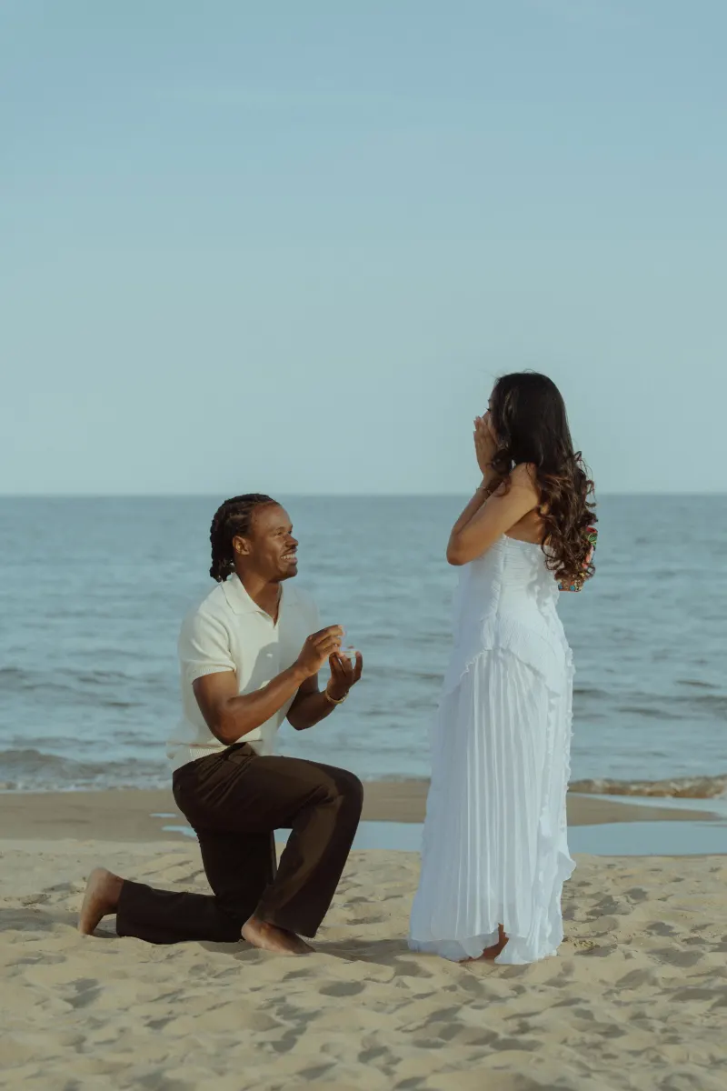 Joyful surprise proposal photoshoot on the beach, capturing pure emotion as he kneels and she reacts in awe.