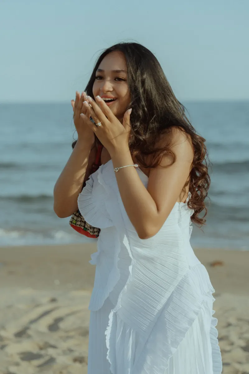 Joyful woman in a white dress reacts to a surprise proposal on the beach, captured during a proposal photoshoot.
