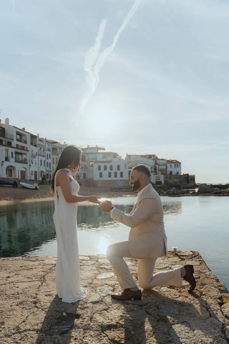 Man proposing on one knee by the water at sunset, capturing a joyful surprise proposal photoshoot moment.