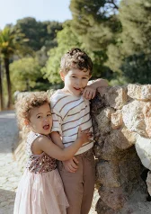 Smiling siblings embrace by a stone wall in the sun, captured during a Vacation Portraits photoshoot in a scenic park.